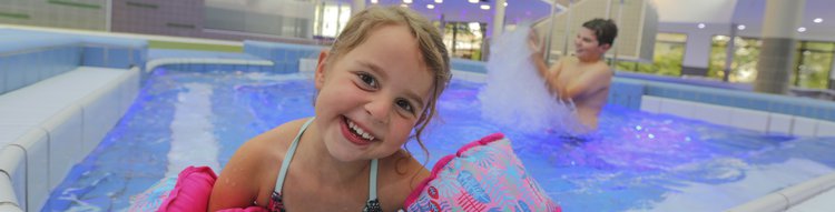 Ein lachendes Kind mit SChwimmflügeln am Beckenrand im Hallenbad