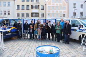 Die beiden Rufbusse stehen auf dem Hauptplatz. Zwischen den Busse stehen drei Schautafeln. Vor den Schautafeln und Bussen stehen 16 Leute, die Teil des Rufbus-Teams sind.