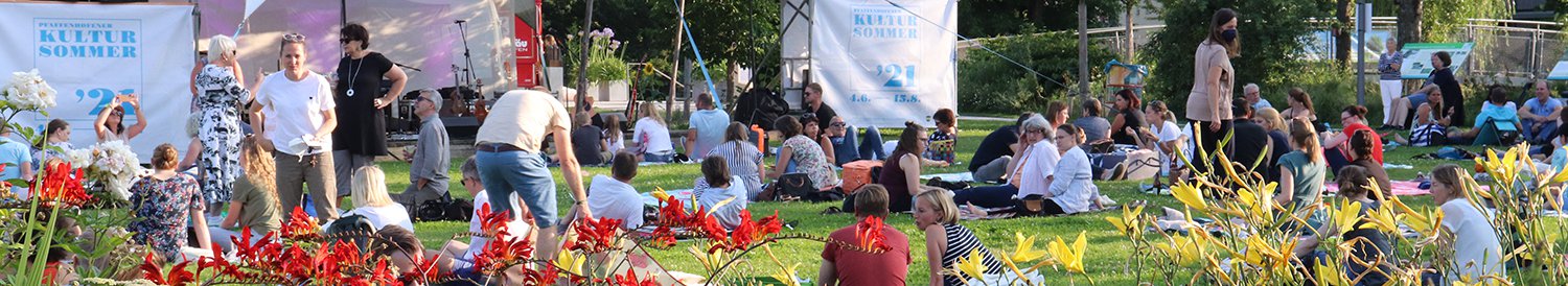 Blumen im Vordergrund, danhinter Menschen auf einer Wiese vor einer Bühne