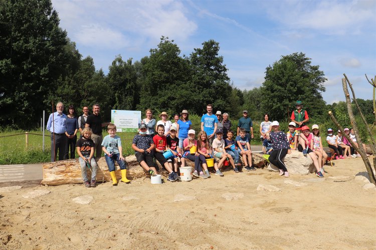 Zahlreiche Erwachsene und Kinder stehen und sitzen hinter einer Sandfläche. Eine Tafel wird hocghgehalten