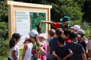 Ein Mann mit Hut erklärt Kindern eine Schautafel auf der ein Lageplan ist