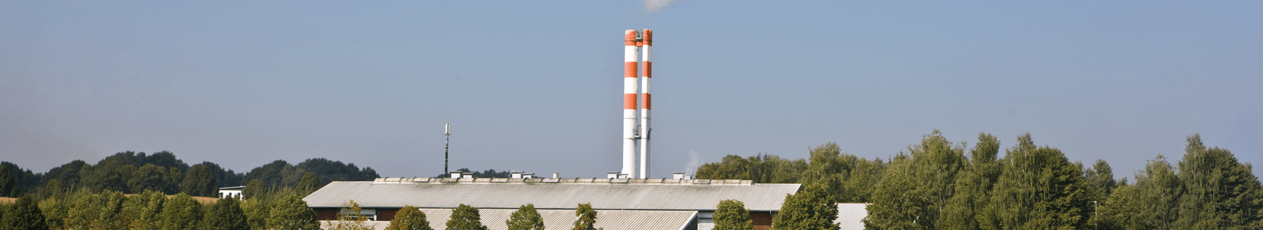 Blockheizkraftwerk Pfaffenhofen: Graues Gebäude mit zwei Rot-weißen Kaminen zwischen grünen Bäumen