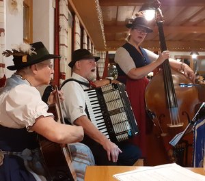Drei Musiker in bayerischer Tracht und mit Instrumenten in einem Wirtshaus.