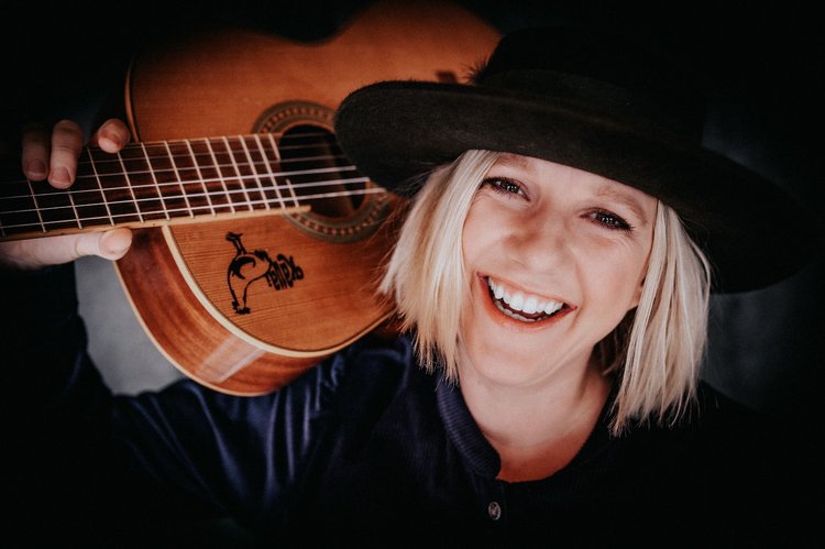 Claudia Koreck mit Hut lacht in die Kamer und hat auf ihrer Schulter ihre Gitarre