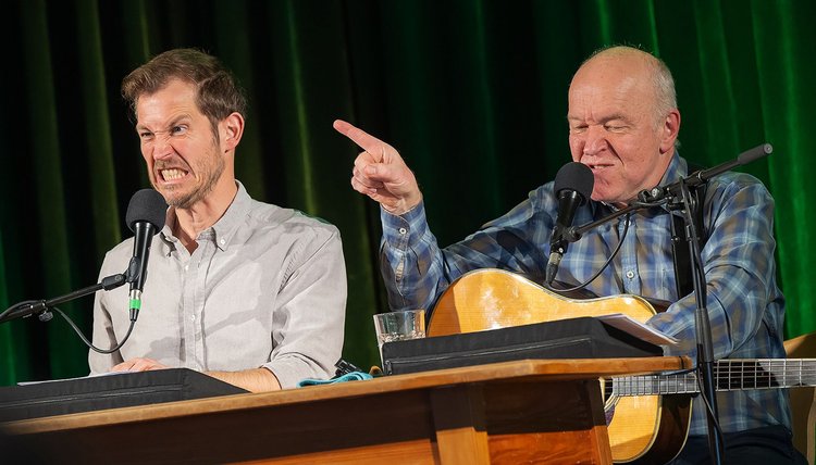 Heinz Josef Braun und Stefan Murr sitzend auf der Bühne an einem Tisch mit einer Gitarre in der Hand